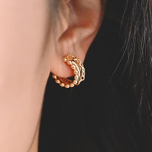 May include: Gold hoop earrings with a beaded design. The earrings are worn in the ear of a person with long dark hair.