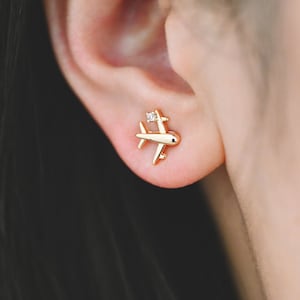 Può includere: Orecchini a lobo in oro a forma di aereo con un piccolo accento di diamante. Gli orecchini sono mostrati nell'orecchio di una persona.