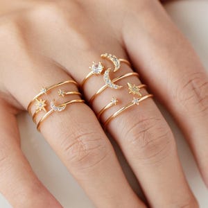 May include: A hand wearing several gold-colored rings. The rings feature celestial designs, including stars and crescent moons, and are adorned with small, clear gemstones. The rings are stacked on the fingers, creating a layered effect.