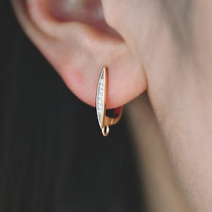 May include: A gold-colored earring with a curved, elongated design. The earring features a row of small, clear gemstones along one side. The earring is attached to a person's ear, showcasing its delicate and elegant appearance.
