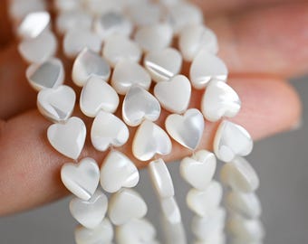 May include: Multiple strands of white heart-shaped beads made from iridescent mother of pearl. Each bead features a smooth, polished surface with a natural pearlescent sheen, arranged in vertical rows to showcase the consistent heart shape.