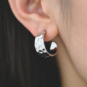 May include: Silver hoop earrings with a hammered texture. The earrings are worn in a person's ear.