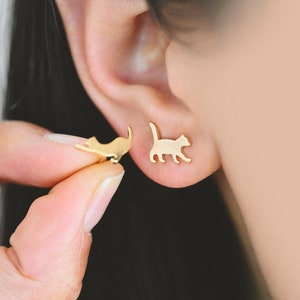 May include: Gold-colored cat-shaped stud earrings. One earring is in the ear, and the other is held by a person's fingers. The earrings are small and minimalist, with a simple silhouette of a cat.