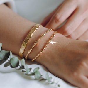 Könnte beinhalten: Ein Satz von vier goldenen Armbändern, die auf dem Handgelenk einer Person übereinander liegen. Die Armbänder sind in verschiedenen Stilen, darunter ein Gliederarmband, ein dünnes Armreif, ein dickerer Armreif mit strukturierter Oberfläche und ein einfaches Stabarmband.