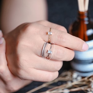 May include: A hand wearing a stack of thin gold and silver rings with a single larger clear stone ring on top. The rings are on the middle finger of the hand.