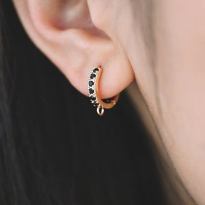 Pode incluir: Brincos de argola dourados com uma fileira de pedras preciosas pretas. Os brincos têm um pequeno charme pendurado.