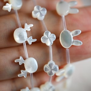 May include: Delicate strands of iridescent white beads, featuring reindeer, flower, and bunny shapes. The beads are strung on clear, thin cords, creating a whimsical design. The beads are held in a hand.