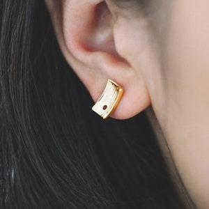 May include: Gold-toned stud earrings with a textured rectangular design. The earrings have a small, round, red stone in the center.