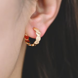 May include: Gold hoop earrings with a textured, slightly rounded design. The earrings are worn in a person's ear.