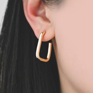 May include: Gold-toned hoop earrings with a rectangular shape. The earrings are worn in a person's ear.