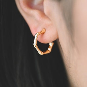 May include: Gold bamboo hoop earrings. The earrings are made of small, round, gold-colored pieces that are connected to form a hoop. The earrings are worn in the earlobe.