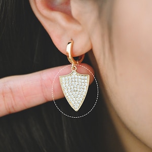 Puede incluir: Aretes de aro dorados con un dije colgante en forma de escudo incrustado con piedras de circonita cúbica transparentes.