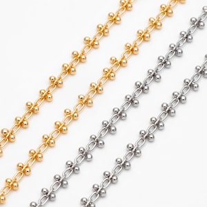 Puede incluir: Dos cadenas, una plateada y otra dorada, con pequeñas cuentas redondas conectadas por un delicado enlace en forma de flor. Las cadenas están dispuestas una al lado de la otra sobre un fondo blanco.