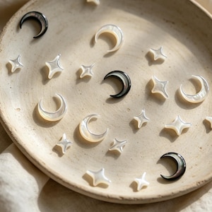May include: A collection of celestial-themed jewelry pieces, including crescent moons and star shapes, arranged on a beige ceramic plate. The moons are black and iridescent, while the stars and moons are pearlescent white.