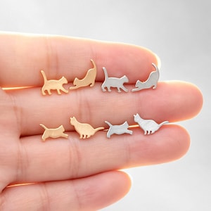 May include: A collection of cat-shaped stud earrings in gold and silver tones. The earrings are arranged in various poses, including walking, stretching, and sitting. The earrings are small and delicate, perfect for cat lovers.