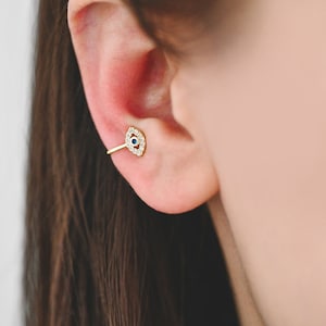 Könnte beinhalten: Goldener Ohrring mit einem kleinen bösen Auge. Der Anhänger ist mit klaren und blauen Edelsteinen besetzt.
