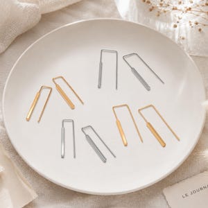 May include: A collection of six U-shaped earrings arranged on a white plate. The set includes three pairs of earrings in gold and silver tones, each featuring a thin wire frame and a solid rectangular bar segment at the base of the U-shape.