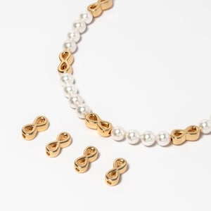 Puede incluir: Un collar de oro con cuentas de perlas blancas y dijes con símbolo de infinito. El collar está colocado sobre una superficie blanca con cuatro dijes de infinito adicionales.