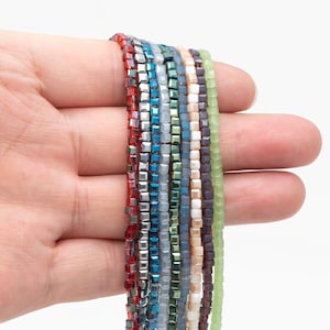 Puede incluir: Una mano sostiene hebras de cuentas cuadradas en varios colores, incluyendo rojo, plateado, azul, verde y morado. Las cuentas son pequeñas y de tamaño uniforme, creando una exhibición texturizada y colorida. Las cuentas son probablemente para hacer joyas.