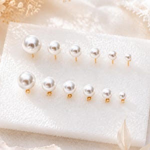 Puede incluir: Una colección de doce pendientes de perlas artificiales blancas esféricas dispuestos en dos filas sobre un bloque de espuma blanca. La fila superior presenta seis pendientes de botón con postes dorados, la inferior seis dijes con aros dorados.