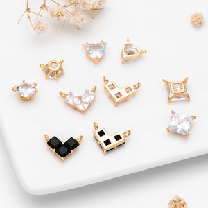 May include: A collection of gold-toned jewelry charms with various shapes, including hearts, squares, and geometric designs. Some charms feature clear or black gemstones, while others have a plain gold finish. The charms are arranged on a white surface.