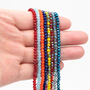 Puede incluir: Una mano sosteniendo hilos de cuentas facetadas de colores. Las cuentas son de varios colores, incluyendo rojo, naranja, amarillo, azul y turquesa. Las cuentas están ensartadas, creando una exhibición vibrante. Ideal para la creación de joyas.