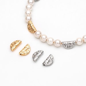 May include: A pearl necklace with gold and silver half-moon shaped charms. The charms have a textured surface. There are also two extra charms on the white surface.