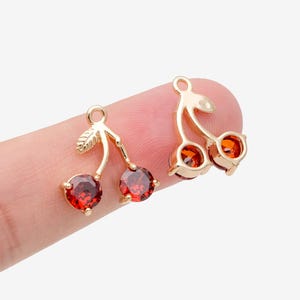May include: Two gold-toned metal cherry charms featuring faceted, round, deep red stones as the fruit and a small leaf detail on the stem. Each charm includes a small circular loop at the top for attachment, showcasing a polished finish.
