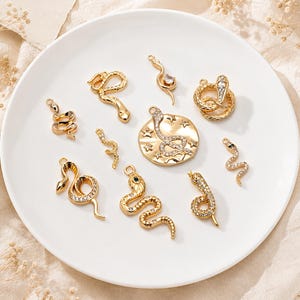 May include: A collection of gold-toned metal jewellery charms featuring various snake designs, some accented with clear crystals and one with a green stone. The charms include coiled snakes, a circular pendant with a snake and stars, and slender snake shapes.