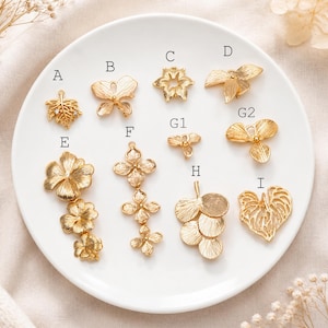 Puede incluir: Una colección de dijes de metal dorado dispuestos sobre un plato blanco, etiquetados de la A a la I. Los dijes presentan diversas formas botánicas y de insectos, incluyendo hojas, flores y mariposas, cada uno con una pequeña argolla.