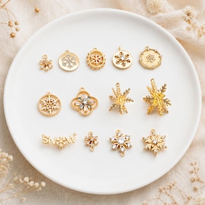 Op de afbeelding: Een collectie goudkleurige metalen bedels met diverse sneeuwvlokontwerpen, sommige versierd met heldere kristallen. De bedels bevatten cirkelvormige, klaverblad- en stervormige motieven, samen met een horizontaal sieraad met het woord SNOW.