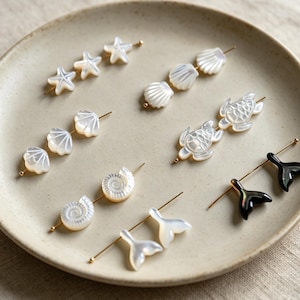 Puede incluir: Una colección de alfileres decorativos con formas talladas en nácar, incluyendo estrellas de mar, conchas, tortugas marinas, caracolas en espiral y colas de ballena, junto a dos alfileres oscuros, dispuestos sobre un plato de cerámica.