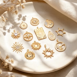 May include: A collection of gold-colored sun-themed charms and pendants. The charms vary in design, including sunbursts, sun faces, and suns with gemstones. They are displayed on a white plate with floral accents, showcasing a celestial jewelry theme.