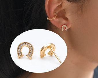 May include: Gold horseshoe earrings with clear cubic zirconia stones. The earrings are shown on a model's ear.