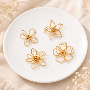 May include: Four gold-toned metal wire ornaments shaped like stylised flowers with open, looping petals. Each piece features a delicate, interconnected wire design that creates a lightweight, airy appearance. The ornaments are arranged on a white plate.