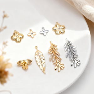 以下が含まれることがあります： ゴールドとシルバーの金属製チャームのコレクション。花の形、金のつぼみ、金の羽、そして金と銀の葉のついた枝のデザインが含まれており、すべてに小さな透明な石が埋め込まれています。