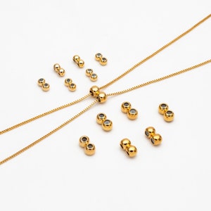 Puede incluir: Un conjunto de cuentas de metal dorado con un centro hueco. Las cuentas están dispuestas en un círculo alrededor de un collar de cadena dorada. El collar es de estilo deslizante con dos hebras de cadena.