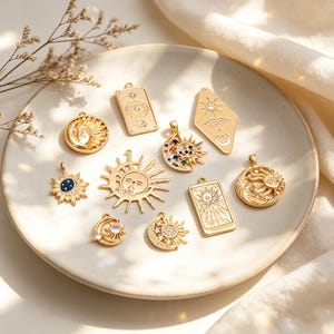Könnte beinhalten: Eine Sammlung goldfarbener Hängeanhänger mit Himmelsmotiven, darunter Sonnen, Monde und Sterne, auf einem weißen Teller angeordnet. Einige Anhänger weisen farbige Edelsteine auf. Die Anhänger haben verschiedene Formen, darunter rund, rechteckig und rautenförmig.