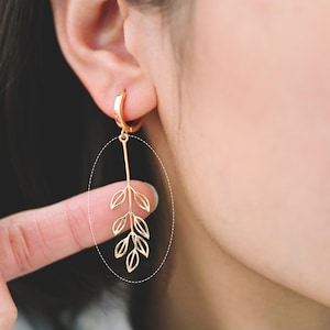 Può includere: Orecchini dorati a forma di foglie. Gli orecchini presentano un piccolo cerchio che si attacca a una barra lunga e sottile. La barra ha una serie di ritagli a forma di foglia. Gli orecchini sono indossati sull'orecchio. Lo sfondo è sfocato.