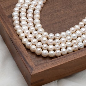 May include: Multiple strands of creamy white pearls, each with a lustrous sheen, arranged in a curved design. The pearls are displayed on a dark brown wooden tray, creating a contrast. The pearls are approximately 0.8 cm in diameter.