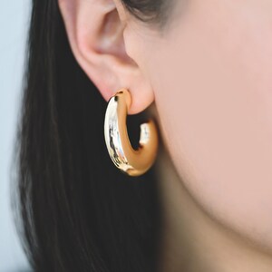May include: A close-up of a gold-colored hoop earring. The earring is thick and rounded, with a smooth, reflective surface. The earring is worn on an ear, with dark hair visible in the background. The earring is a bold, modern jewelry piece.