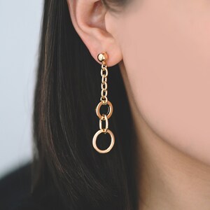 Puede incluir: Pendientes colgantes dorados con un pequeño perno de bola, una cadena y tres colgantes circulares conectados. Los pendientes se llevan en la oreja de una persona, con su pelo oscuro y su hombro visibles en el fondo.