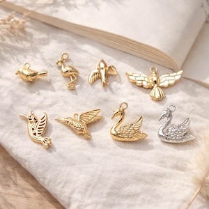 May include: A collection of eight small metal charms in the shapes of various birds, including swans, hummingbirds, and other avian figures. Seven charms are gold-toned, and one swan charm is silver-toned, each featuring a small loop for attachment.