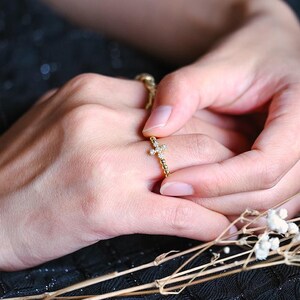 May include: A pair of hands holding a gold ring with a small diamond. The hands are resting on a black fabric with a bouquet of white flowers and a brown glass bottle with wooden sticks.
