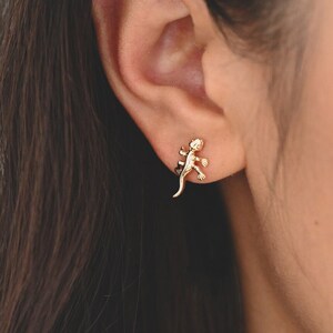 May include: A close-up of a person's ear with a gold lizard stud earring. The earring is small and delicate, and the lizard is facing forward.