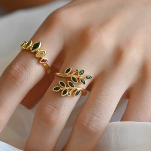 May include: Two gold rings with gemstone accents. One ring features a row of black and red gemstones, while the other has a leaf design with black gemstones. The rings are displayed on a hand against a white background.
