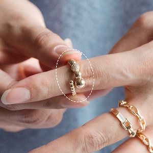 May include: A gold ring with a textured design and a large round bead. The ring is worn on a finger with other gold rings.