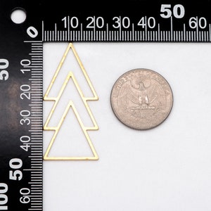 May include: A gold-colored metal charm in the shape of a stylized tree, next to a quarter coin for size comparison. The charm is composed of three connected triangles. The background is white with a black ruler along the left side.
