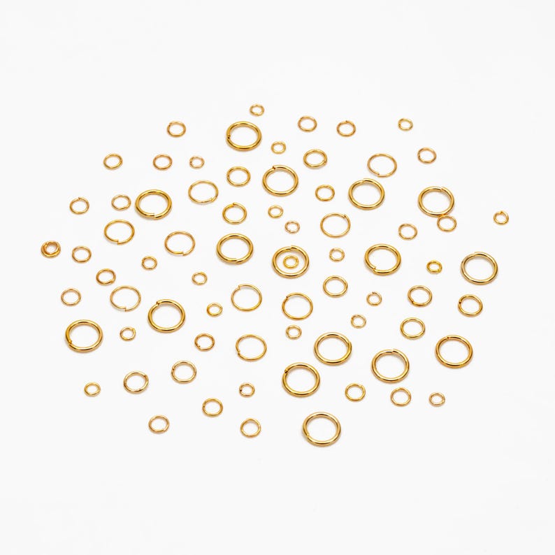May include: A collection of gold jump rings of various sizes scattered on a white background.