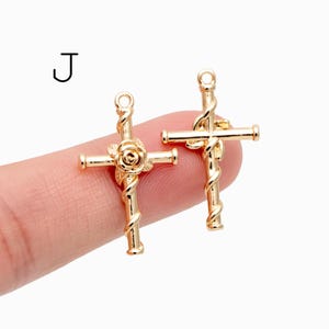 Pu&ograve; includere: Due ciondoli a forma di croce color oro. Una croce presenta un design a rosa, mentre l'altra ha un dettaglio a vite intrecciata. Entrambe le croci hanno un piccolo anello nella parte superiore per essere appese. La lettera "J" &egrave; nell'angolo in alto a sinistra.